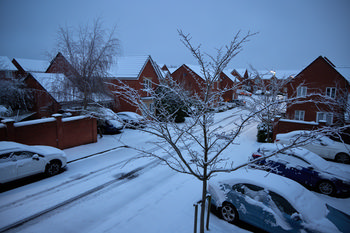 Suburban snow This urban landscape photograph depicts a suburban street in Derbyshire, England, in the United Kingdom, blanketed by fresh snow. Captured in the early winter morning, but with lighting conditions typical of evening, the scene shows parked cars and red-brick houses characteristic of the area, all covered in snow. Leafless trees line the street, their branches outlined in white, contributing to the winter atmosphere. The photograph highlights the peacefulness of a typical residential neighborhood in Derbyshire during the snowy winter season.